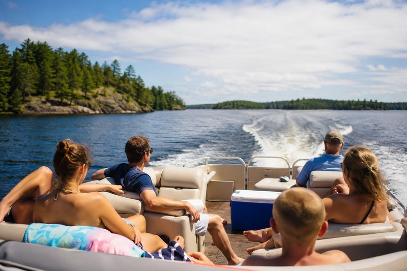 Friends relaxing on a Lazy Loon pontoon boat cruising past forested islands on a sunny Northwoods lake.